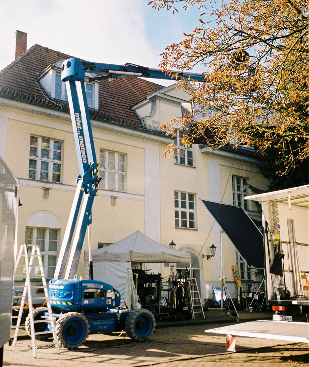 An outdoor film set is visible with a blue boom lift extended towards a light-colored building, alongside a white tent and film equipment. An autumn tree branch is in the upper right. KLEINANZEIGEN by Arrigo Reuss for Kleinanzeigen — © EASYdoesit.