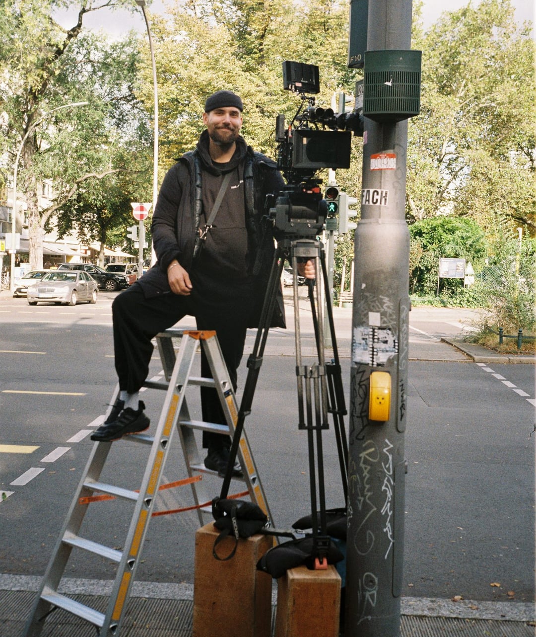 A smiling man on a ladder operates a professional camera on a tripod at a street intersection with trees and cars in the background. KLEINANZEIGEN by Arrigo Reuss for Kleinanzeigen — © EASYdoesit.