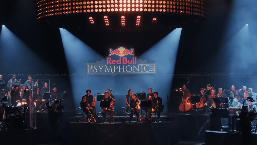 An orchestra performs on a stage under bright spotlights and a large circular light fixture. REDBULL SYMPHONIC by Chris Schwarz for RedBull — © EASYdoesit.