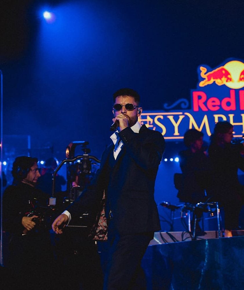 A man in a suit and sunglasses sings into a microphone on a blue-lit stage. A camera operator is visible on the left, with other figures in the background. REDBULL SYMPHONIC by Chris Schwarz for RedBull — © EASYdoesit.