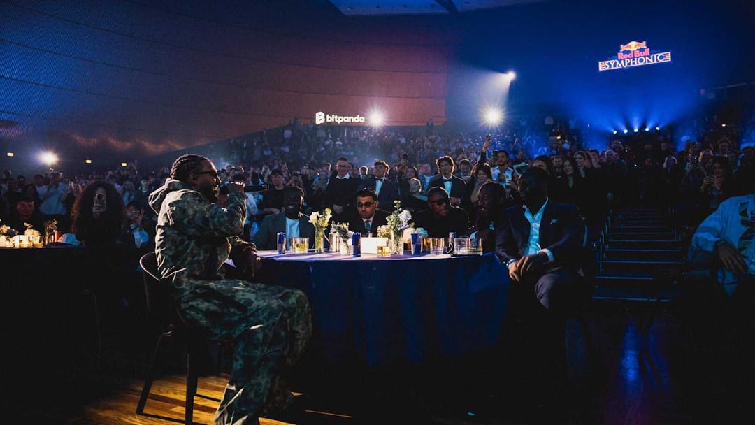 A person in camouflage clothing speaks into a microphone while seated on a chair, facing an audience in a large, dimly lit venue. Several people are seated at a table in front of the speaker, with a large crowd visible in the background under blue and red stage lights. REDBULL SYMPHONIC von Chris Schwarz für RedBull — © EASYdoesit.