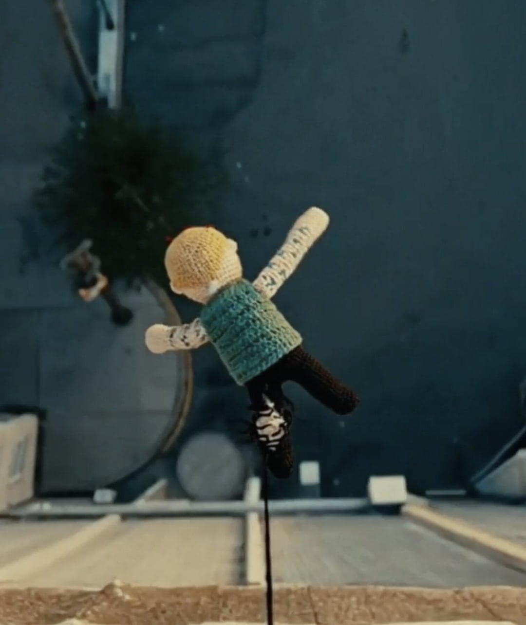 A knitted doll with a yellow head and teal vest hangs or falls against a dark background, viewed from above. KONTRA K – TRAILER von Christoph Szulecki für Kontra K — © EASYdoesit.