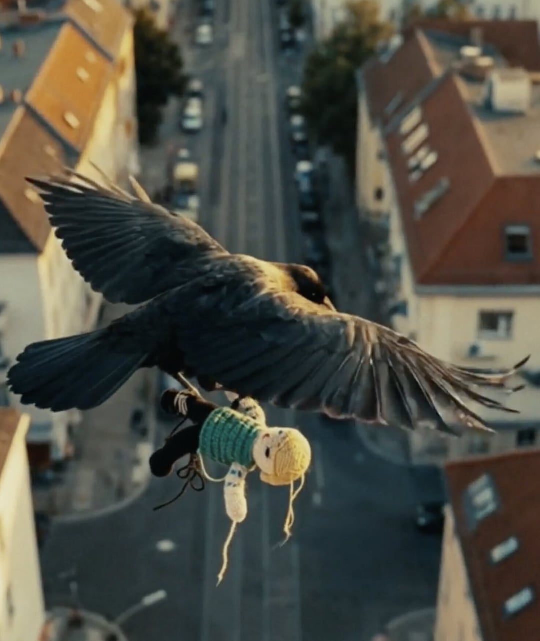 A large dark bird flies high above a city street, carrying a knitted doll. KONTRA K – TRAILER by Christoph Szulecki for Kontra K — © EASYdoesit.