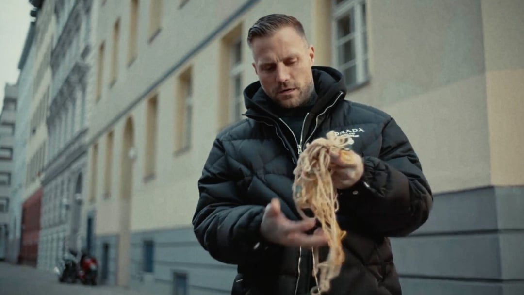 A man in a black puffer jacket stands on a street, looking down at a tangled string he holds in his hands. Buildings line the street in the background. KONTRA K – TRAILER by Christoph Szulecki for Kontra K — © EASYdoesit.