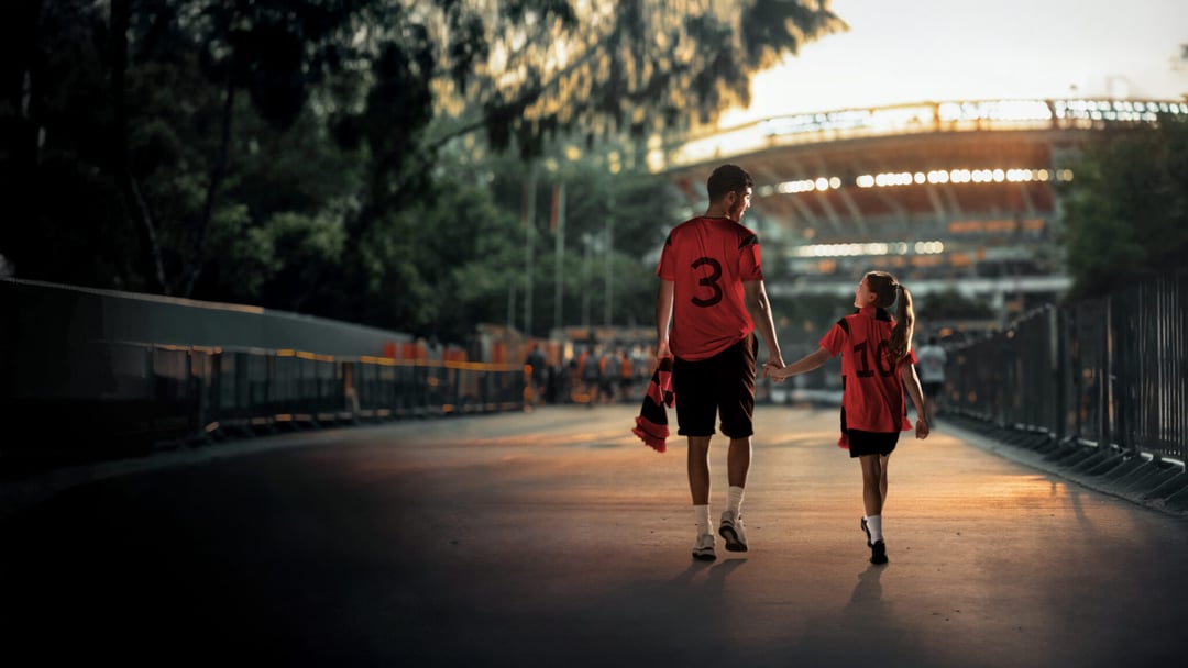 A man and a young girl, both in red jerseys, walk hand-in-hand away from the viewer on a path, with a large stadium visible in the background under a warm sky. MASTERCARD von Luca Homolka für Mastercard — © EASYdoesit.